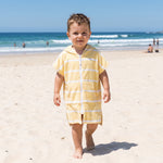 Toddler boy wearing Amalfi yellow and white striped toddler hooded towel, on bright dry beach sand