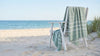 Sage green Turkish towel with white stripes and hand-twisted tassels draped over a white timber beach chair on the sand, with the sea behind