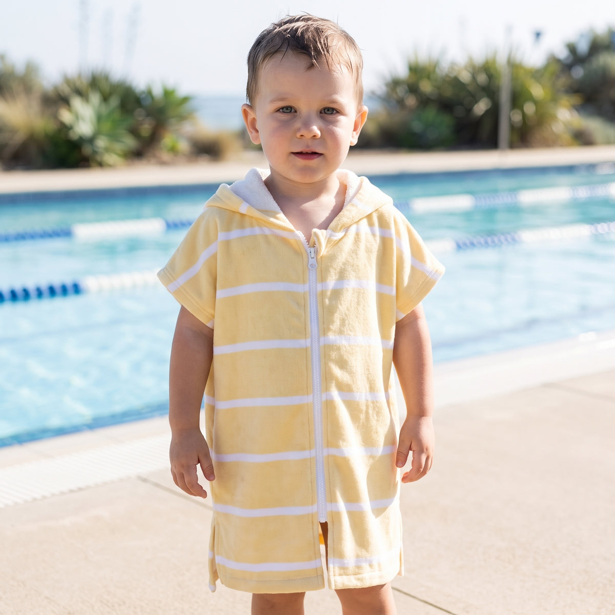 Amalfi toddler hooded towel in yellow and white stripes worn by a toddler boy, beside an outdoor pool