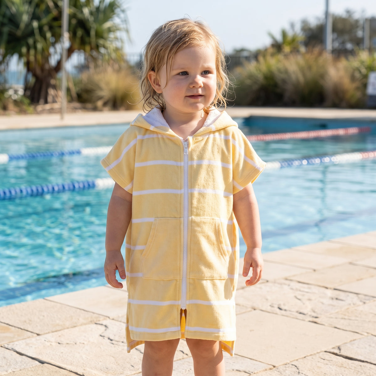 Toddler girl wearing Amalfi yellow and white striped toddler hooded towel, beside an outdoor pool